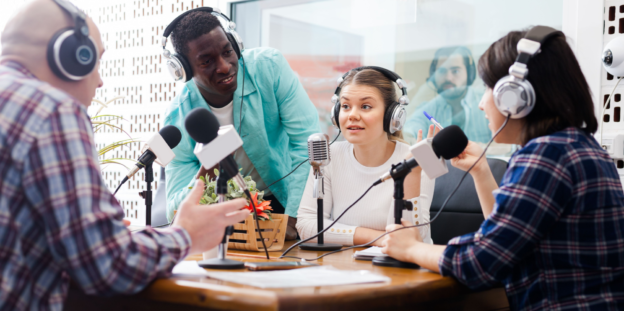 Group of people wearing headphones sit around a table with microphones on it