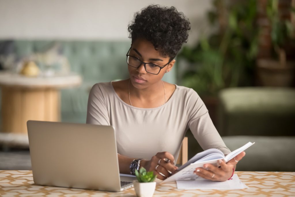 Woman sits at table in front of laptop with notepad