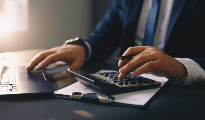 A person's hands on a calculator and a laptop on a table