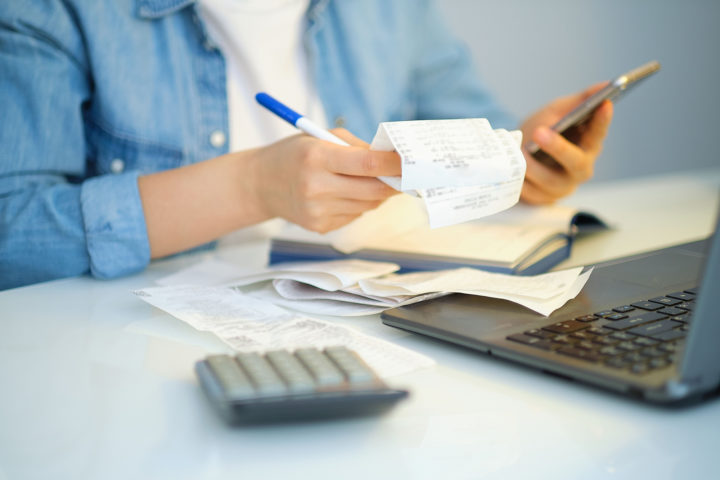 A person's hands holding a pen, receipts and a mobile phone in front of a laptop and calculator