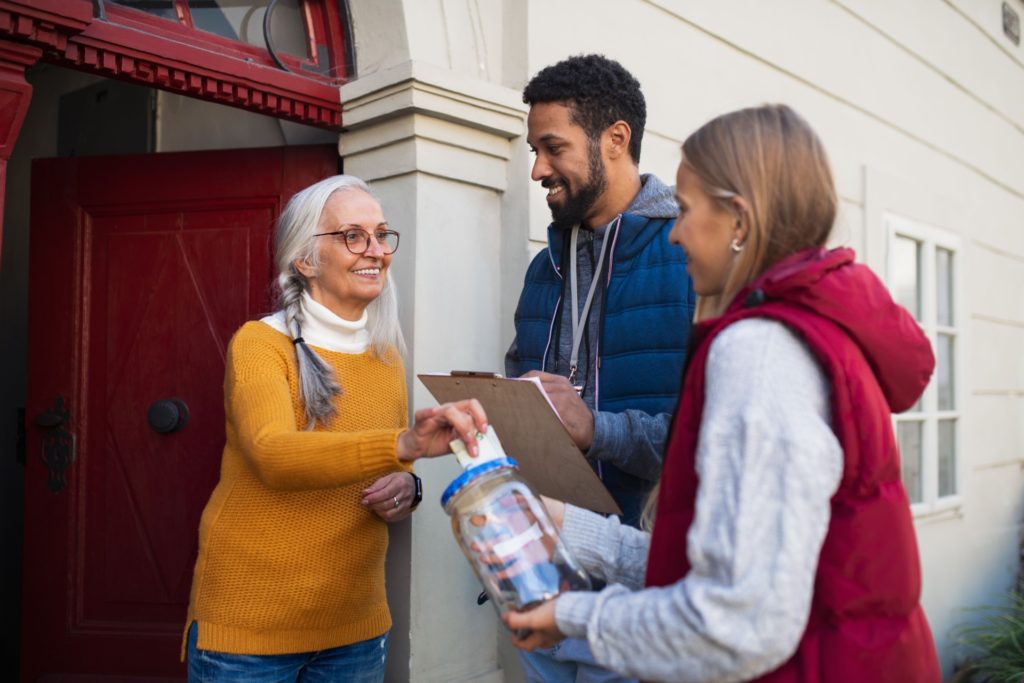 Volunteers collecting donations at a doorstep, speaking with a resident and holding a collection container