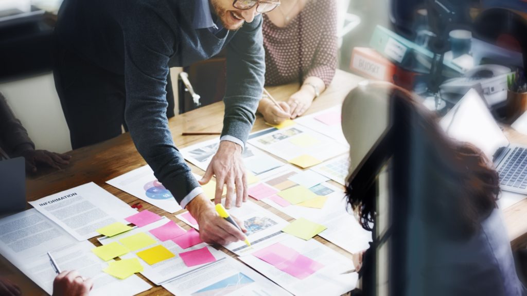 People stand around table writing on pieces of paper covered in post-it notes