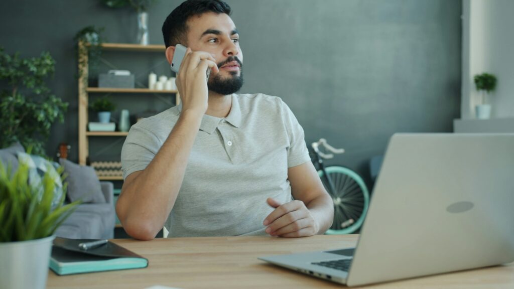 Man talking on a mobile phone while sitting at a desk with a laptop