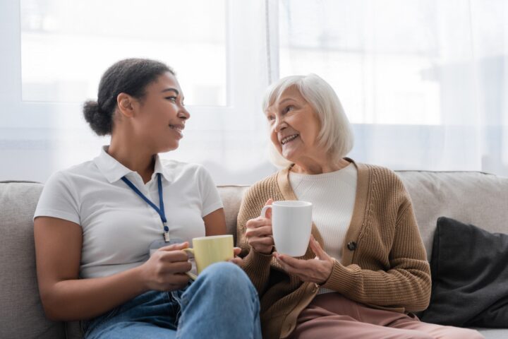 Older woman and younger woman sat on couch talking over cups of tea